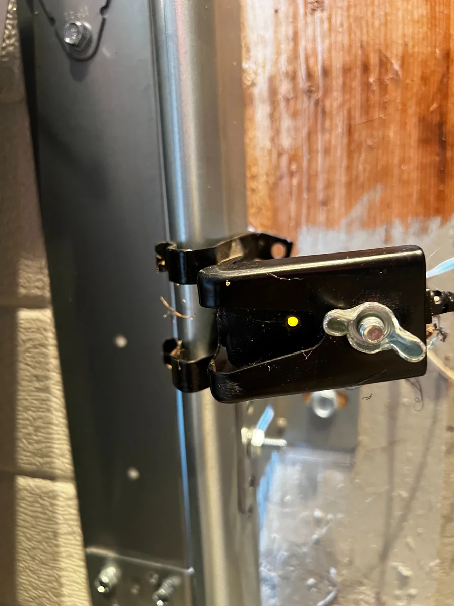 Garage door safety sensor mounted beside the track with its indicator light illuminated.